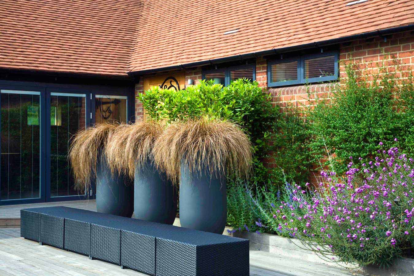 Specimen grasses in three tall planters next to a seat by the stable buildings. Rae Wilkinson Garden and Landscape Design - Garden Designer Sussex, Surrey, London, South-East England